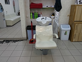 стопка чистых полотенец, шампуни, бальзамы во флаконах с дозатором на полках встроенной ниши в стене, белое кресло с мойкой, контейнеры для мусора в светлом просторном салоне парикмахерской