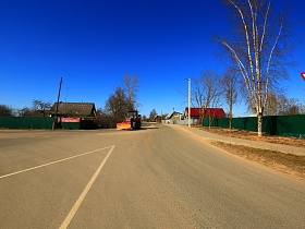 трактор на ровной гладкой асфальтированной дороге в городке Сычево с пешеходной дорожкой вдоль жилых домов за высоким зеленым забором