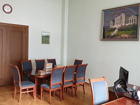 деревянные стулья со спинкой с мягкой серой обивкой вокруг овального стола для заседаний в углу рабочего кабинета КГБ СССР с картиной на стене и круглыми часами над входной дверью для аренды для съемок кино