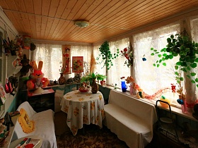 скатерть на круглом столе и покрывала на двух диванах в светлой крытой веранде с многочисленными мелочами-игрушками, вазами,пано, искуственными цветами на больших окнах с гардинами в избе в деревне