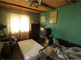 белый матрас на полу, серый мягкий разложенный диван, телевизор на тумбе в спальной комнате с деревянным потолком и зелеными полосатыми обоями