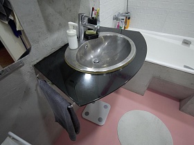 раковина на темной стеклянной поверхности с полотенцами по бокам, белая ванна с экраном, круглый коврик на розовом полу светлой ванной комнаты стильной трешки