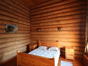 деревянная кровать и прикроватные тумбочки у бревенчатых стен с картиной и бра в комнате отдыха со светлой шторой на окне