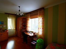 детская корзина с игрушками в виде зеленой лягушки, сиреневый детский стульчик у окна с желто-коричневой гардиной,деревянный шкафчик с игрушками у стены с детскими рисунками на зеленой стене детской комнаты трехкомнатной квартиры