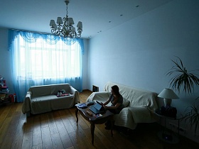 коричневый журнальный столик на полу белой гостиной с двумя мягкими диванами, голубой гардиной на большом окне и подвесной люстрой на потолке современной квартиры в красном кирпичном доме