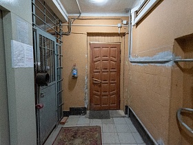 цветной коврик на полу с квадратной плиткой у металлических дверей с решеткой на крышу сталинского дома в светлом освещенном подъезде с бежевыми стенами и белым потолком
