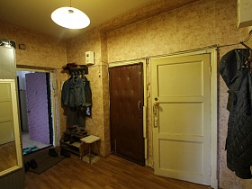 старый  табурет, обувная полка под настенной вешалкой с одеждой, обувь на коврике у открытой входной двери в прихожую простой квартиры в жилом доме