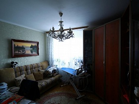 картина над полосатым мягким диваном с подушками, гладильная доска и угловой шкаф для одежды в комнате большой квартиры врача