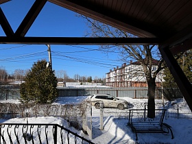 деревянные качели в снежном сугробе у расчищенной дорожки к открытым входным воротам на участок загородного дома