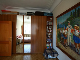чемоданы и коробки на коричневом шкафу для одежды с зеркальными дверцами по центру, большая картина с изображением Гагарина в окружении пионеров на стене спальной комнаты стильной квартиры художника