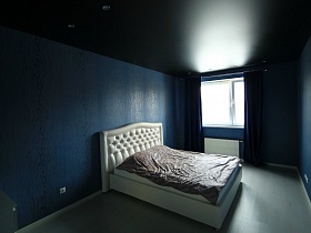просторная синяя спальная комната с черным потолком, большой белой кроватью с мягкой спинкой на светлом полу и темно синими шторами на окне молодежной квартиры