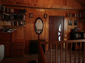 полочки с книгами, овальное зеркало в рамке на стене деревянного холла на лестничной площадке с комодами,столиком и этажеркой  классической семейной дачи