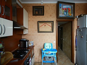 детское бирюзовое кресло для кормления ребенка у стены с картинами, коричнево- белая мебель, белый холодильник в углу кухни семейной трехкомнатной квартиры времен СССР