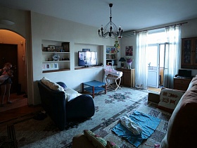 шкафчик с настольной лампой и цветами, детская люлька, журнальный голубой столик у стены с нишами под мебель в гостиной семейной трешки