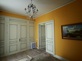 белый встроенный шкаф на всю стену, люстра, стилизованная под свечи над большой кроватью, картина на желтой стене спальни девчачьей современной квартиры с видом на Москву реку