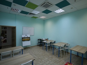 учебная доска на голубой стене, большие деревянные счеты на белом планшете у стола воспитателя напротив деревянных парт классной комнаты детского центра