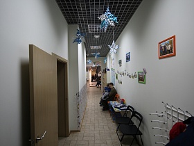 черные стулья для посетителей, детский яркий столик у белых стен с фотографиями светлого коридора с объемными бумажными фонарями, свисающими с потолка с черной мозаичной плиткой