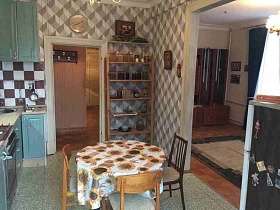 стулья со спинкой вокруг круглого стола с цветочной скатертью в центре кухни с серой плиткой на полу, круглыми часами над открытой дверью и деревянной этажеркой с баночками в углу кв 27
