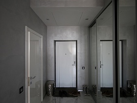 обувь на коврике и серебристый металлический стульчик на полу у белой входной двери в прихожей с большим шкафом-купе у серого цвета стены современной лаконичной минималистической квартиры