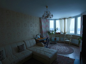 мягкий светлый диван с квадратными подушечками, ковры на полу гостиной с большим эркерным окном, бежевыми стенами и подвесной люстрой на белом потолке трехкомнатной квартиры в Химках