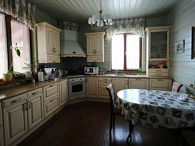 стулья вокруг круглого стола с клеенкой на белой кухне с мебелью молочного цвета просторного современного деревянного дома
