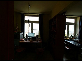 шкаф с книгами на открытых полках между обеденным столом и рабочим в зонированной комнате двухкомнатной неприбранной квартиры с видом на Москва-сити