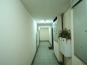 белый потолок, стены, квадратная плитка на полу длинного коридора на первом этаже с входными дверьми в жилые квартиры