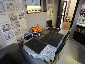 апельсины в вазе на обеденном столе с серыми салфетками на белой ажурной скатерти, стулья со спинкой, серое кресло у стены с зеркалом и декоративной отделкой кухни дизайнерской трешки