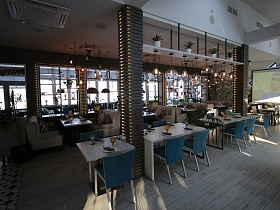комнатные цветы в белых горшочках на подвесной белой полке с подвесными прозрачными светильниками над белыми сервированными столиками между подсвеченными колонами стильного ресторана в светлом стиле