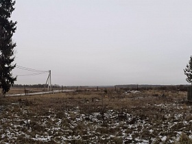вид на открытое поле с засохшей травой, столбиками забора и столбами с электропроводами вдоль дороги в деревне