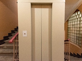 кремовая лифтовая кабина в подъезде сталинского дома с коричневыми стенами над лестничными пролетами с перилами
