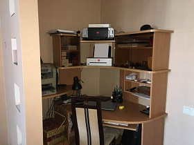 стул с высокой спинкой у компьютерного стола с ноутбуком, канцерярскими принадлежностями, настольной лампой на поверхности, с принтером, сканером, на полках в зонированной спальной комнате