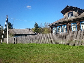 деревянный дом с голубыми наличниками, мансардой за деревянным штакетным забором в старой деревне