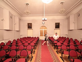 общий вид светлого актового зала эпохи СССР с красной дорожкой между  рядами красных секционных стульев на паркетном полу, большими портретными картинами и круглыми часами над входной арочной дверью