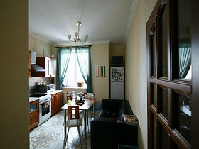 коричневая мебель,черный диван, белый холодильник в светлой кухне с голубой гардиной на окне через открытую дверь трехкомнатной квартиры в жилом доме