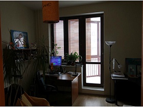 комнатные цветы, кресло, большая рамка с фотографией на настенной полке, компьютерный стол у окна и торшер в стиле хай-тек в рабочей комнате простой квартиры для съемок кино