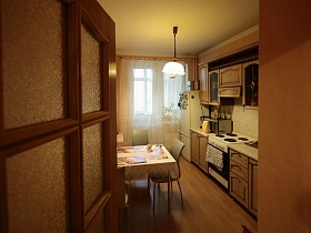 обеденный стол у стены, газовая плита,мойка в бежевой мебельной кухне с коричневым линолеумом на полу современной квартиры молодоженов сквозь открытую дверь