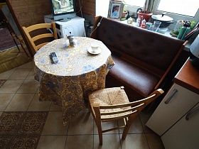 деревянные стулья со спинкой у круглого обеденного стола с цветной клеенкой , коричневый диван у окна на полу кухни с бежево коричневой плиткой трехкомнатной квартиры времен СССР