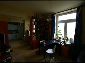 черный стол с настольной лампой, комод у окна с комнатными цветами на подоконнике зонированной комнаты двухкомнатной неприбранной квартиры с видом на Москва-сити