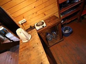 этажерка с посудой, мусорное ведро у тумбы с электроплитой и электочайник на деревянном столе в зоне кухни лачуги отшельника у маленького озера