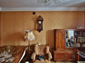 книжный шкаф, мягкая игрушка и сумка на бежевом мягком кресле, торшер и большая кровать с коричневым покрывалом у персиковой стены с деревянными настенными часами с маятником в спальне квартиры эпохи СССР