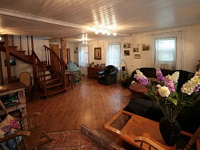 общий вид уютной светлой гостиной с высокими ветками сирени в вазе на столе,деревянной лестницей с резными перилами на второй этаж классической семейной дачи