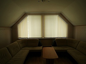 угловые диваны у трапецевидного окна с закрытыми вертикальными жалюзи в светлой гостиной съемного дома в сосновом лесу