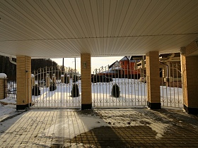 просторная площадка под навесом с кирпичными колонами за белым металлическим забором вокруг участка современного кирпичного коттеджа