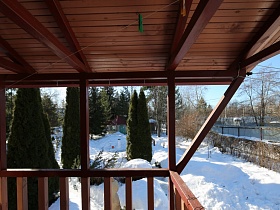 открытая деревянная терраса с перилами оригинального загородного дома с высокими зелеными кипарисами на заснеженном участке