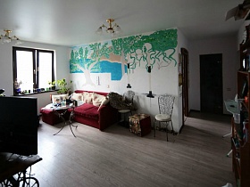 журнальный стол у бардового углового дивана, пуфик и два стула с резной спинкой у стены с рисунком в гостиной простой трехкомнатной квартиры