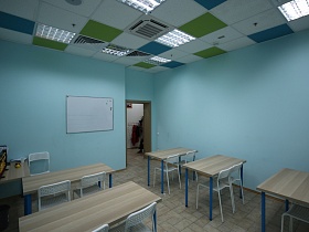 открытая дверь в учебный класс с партами у голубых стен, синими и зелеными плитами на белом потолке с кондиционером и ярким освещением