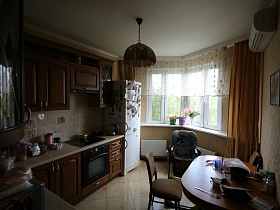 белый холодильник с магнитами на дверце у окна с комнатными цветами в кухне обычной квартиры в новострое
