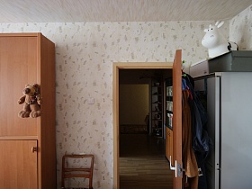 игрушка белой пятнистой коровы, картонные коробки на верху белого шкафа за открытой дверью с домашней одеждой на крючках спальной комнаты современной квартиры неработающих пенсионеров, проживающих с внуками