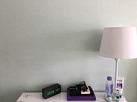 черные электронные часы, очки, телефоны и планшет, косметические средства на поверхности настольной лампы на белом комоде в бирюзовой спальной комнате современной квартиры молодоженов